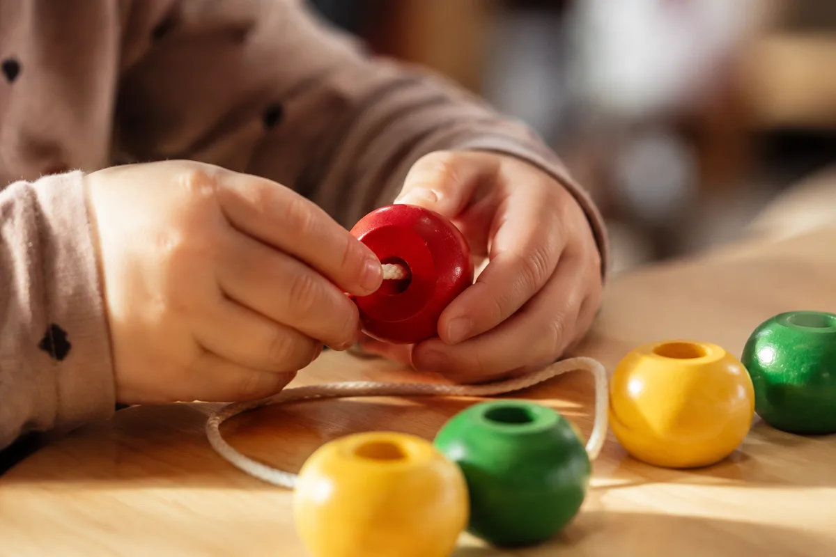 Mains d’enfant enfilant de grosses perles en bois colorées sur un lacet, table basse en bois clair, lumière naturelle douce