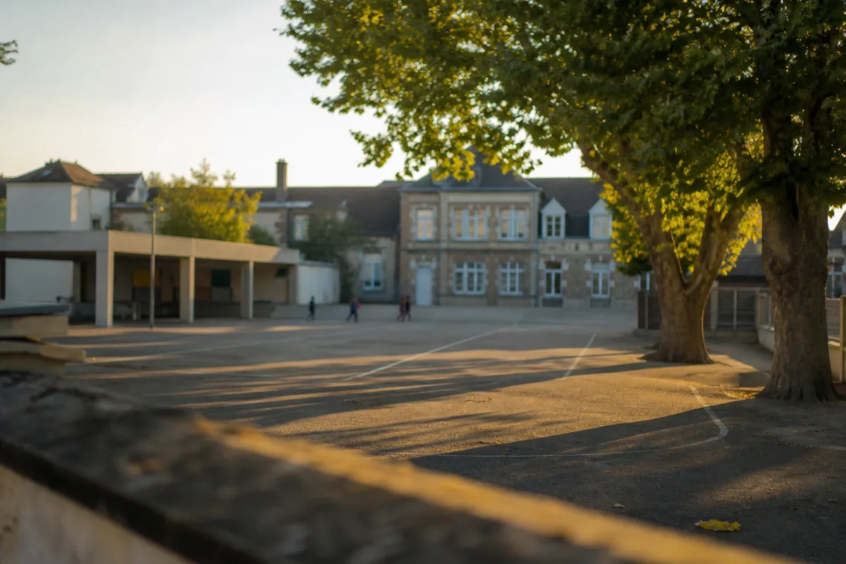 Cour d&rsquo;école française vue de loin en fin d&rsquo;après-midi, silhouettes floues sous le préau